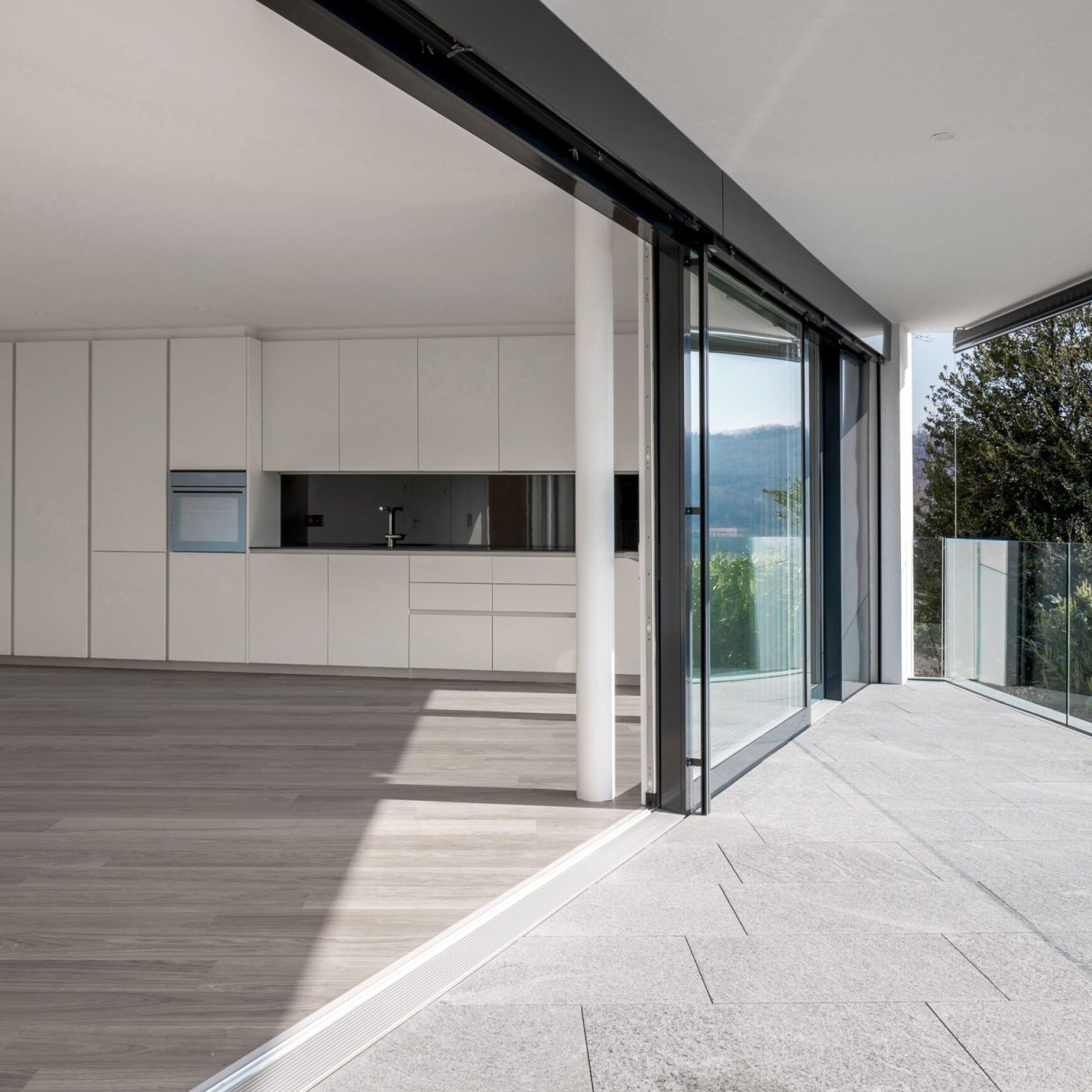 Large empty dining room with kitchen attached to the wall and wooden floor. The space is empty and the adjacent terrace is triangular. It is modern architecture and the window is open.